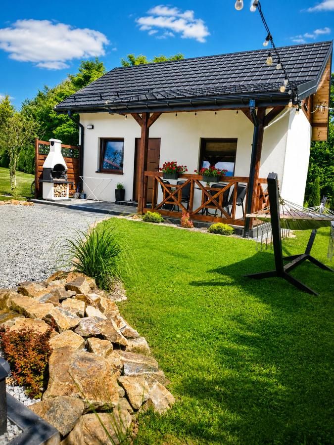 Dom z zadaszonym tarasem i kwiatami, obok grill i hamak na trawniku